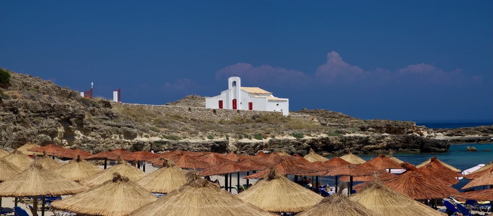 Thatched sunshades on Agios Nikolaos beach in Vasilikos, with the white chapel of St Nicholas on the rocks behind