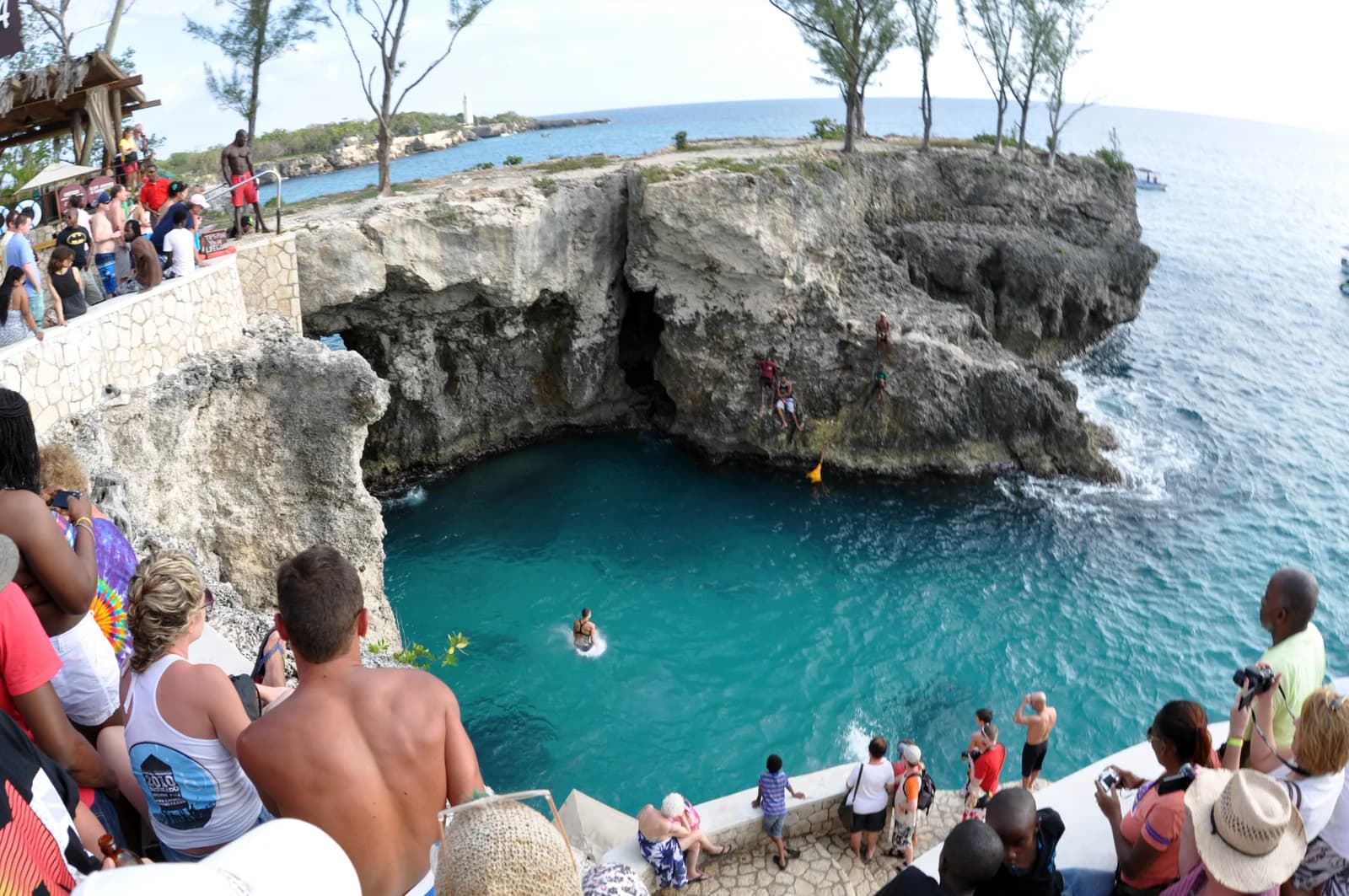 A cliff jumper above the deep turquoise cove at Rick's Cafe on Negril's West End cliffs, with a crowd watching from the rocks