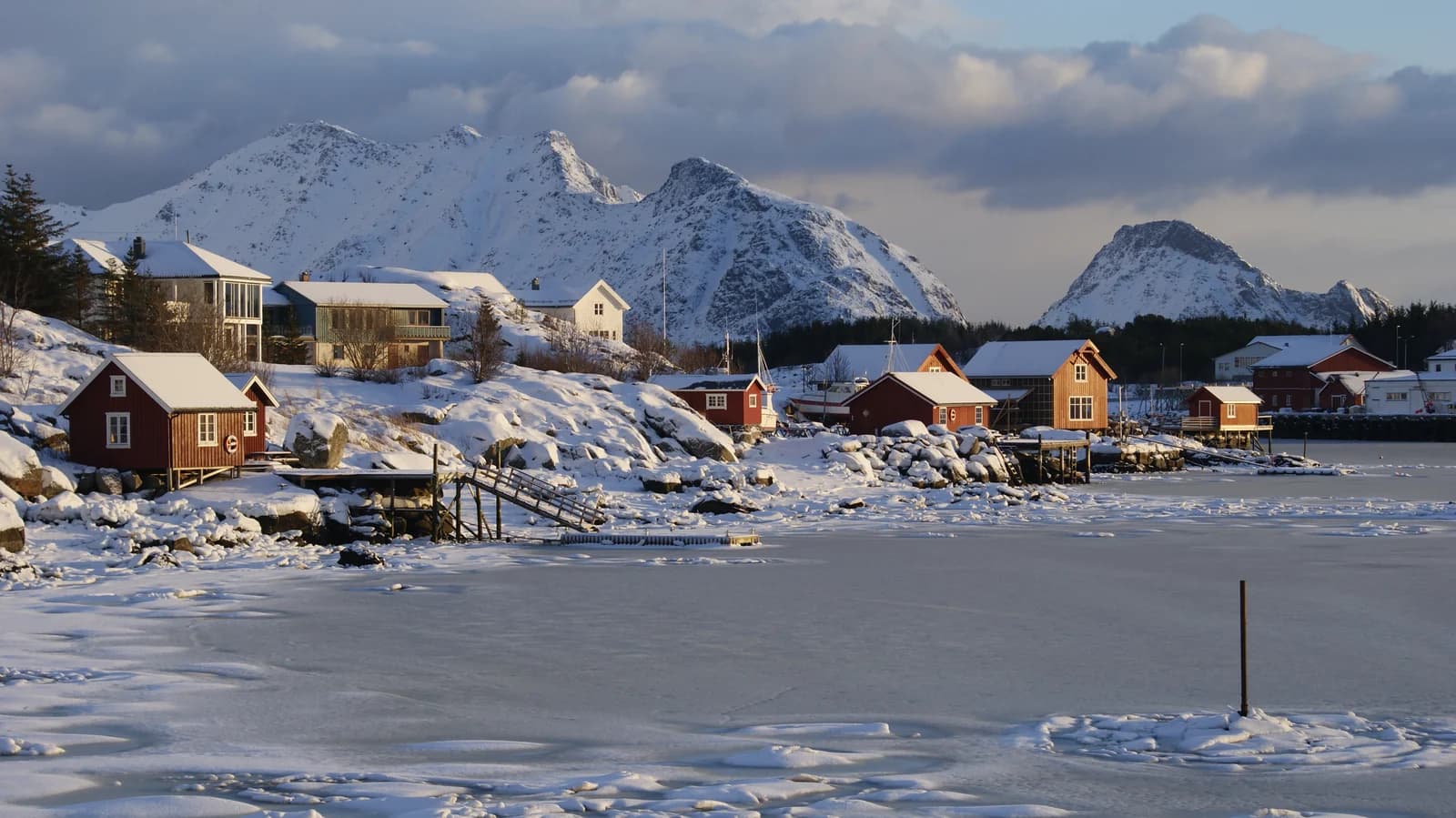 Snow covered wooden cabins along the icy shore at Ballstad in Lofoten below sharp winter peaks