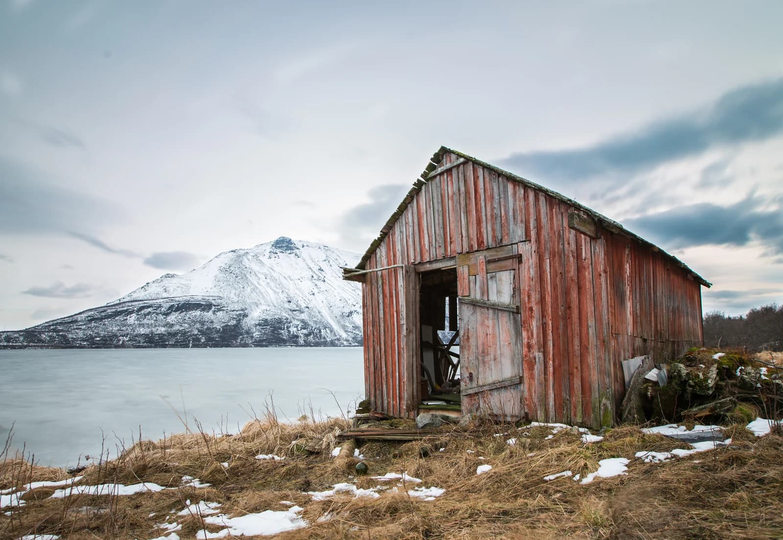 A red boathouse on the fjord shore with the snow covered Lyngen Alps across the water