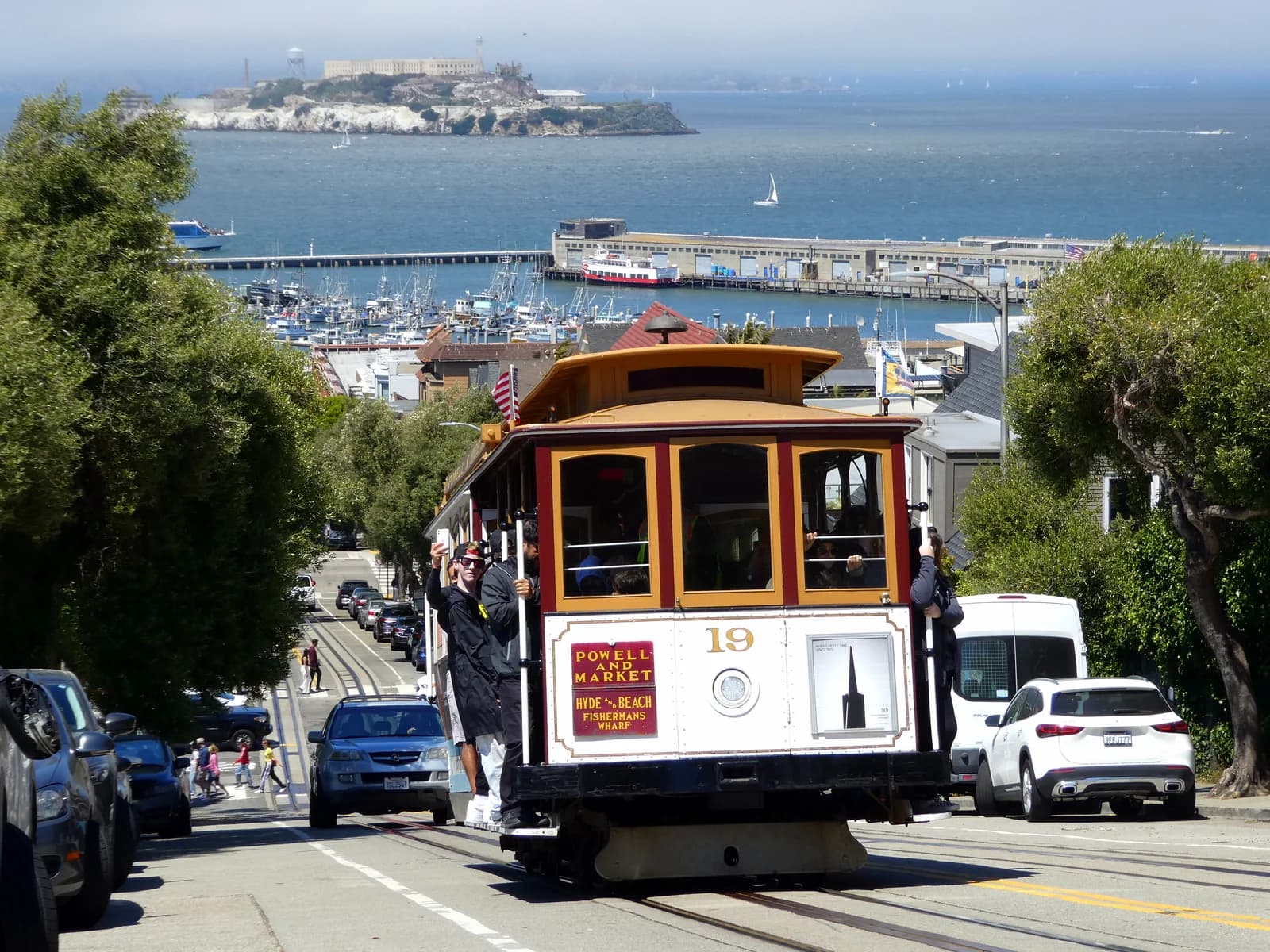 Riders hang off a Powell-Hyde cable car as it crests Hyde Street with the bay and Alcatraz behind