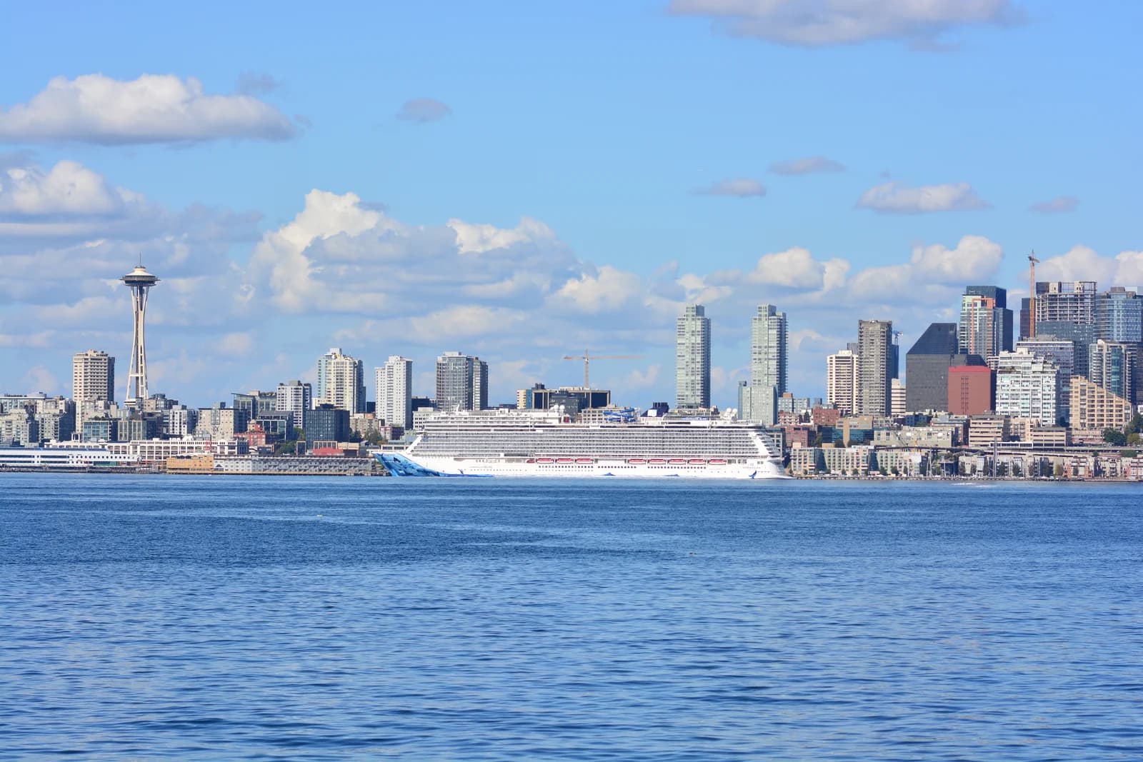 A cruise ship docked at the downtown Seattle waterfront, with the Space Needle and the skyline behind it across Elliott Bay