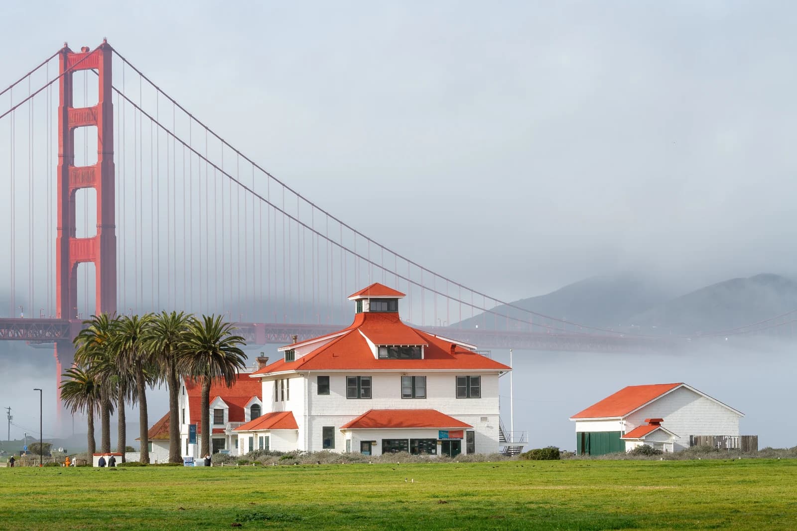 Fog pours over the Golden Gate Bridge behind the red-roofed old Coast Guard station at Crissy Field