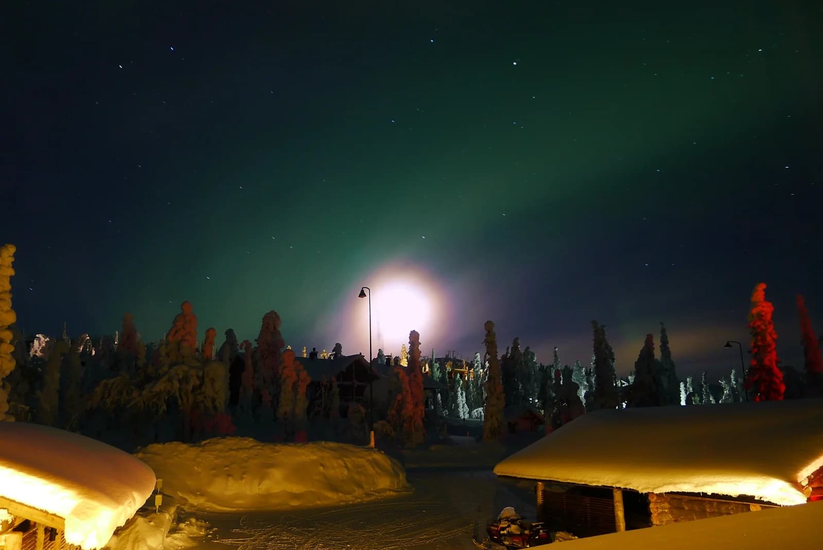 A green aurora arc and a bright moon in the same night sky over snow-covered forest at Ruka in Finnish Lapland