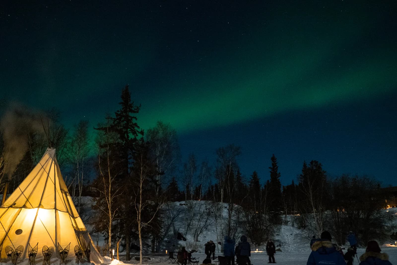Green northern lights above a lit teepee and snowy forest at Yellowknife in the Northwest Territories