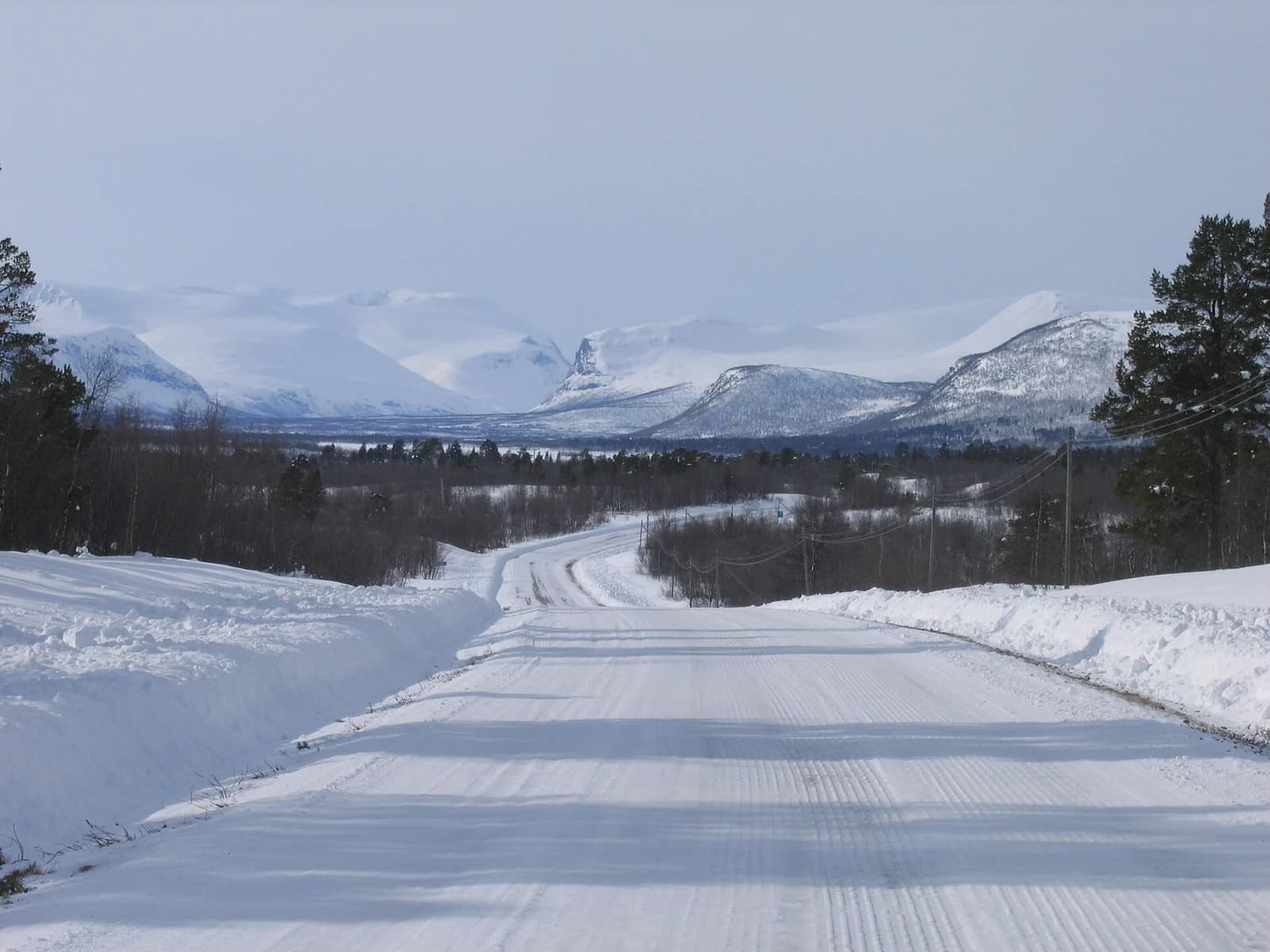 A snow-banked road running through birch forest and open fell in Swedish Lapland in late winter