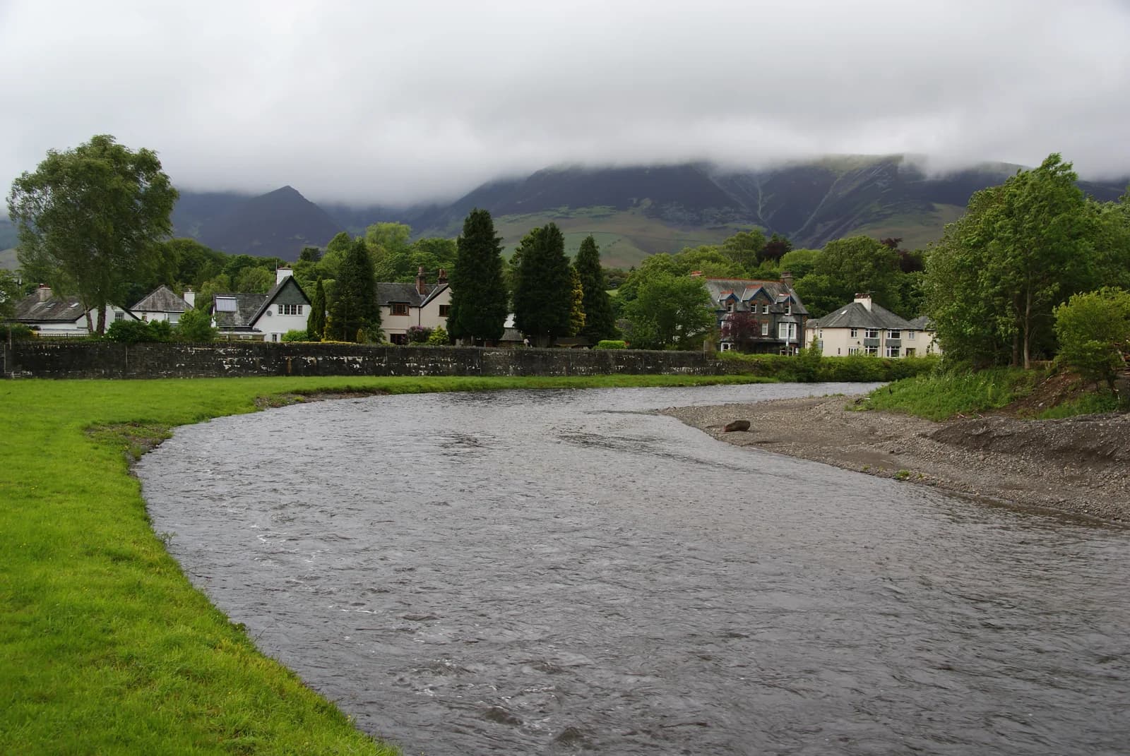The River Greta curving past houses and a riverside green in Keswick, with cloud on the fells