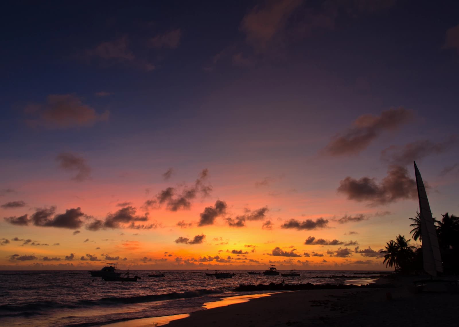 Sunset over a Barbados beach, with fishing boats at anchor, a catamaran drawn up on the sand and palms in silhouette