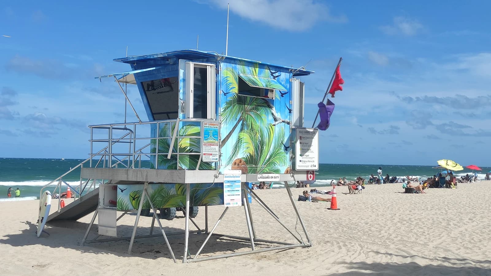 A painted Fort Lauderdale lifeguard tower flying warning flags, with a beach warning sign board beside the steps
