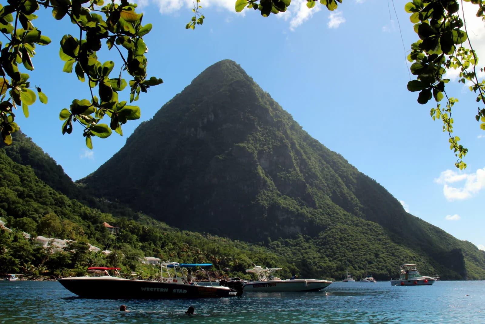 A Piton rising above the bay at Soufriere in Saint Lucia, framed by sea grape leaves with boats moored below