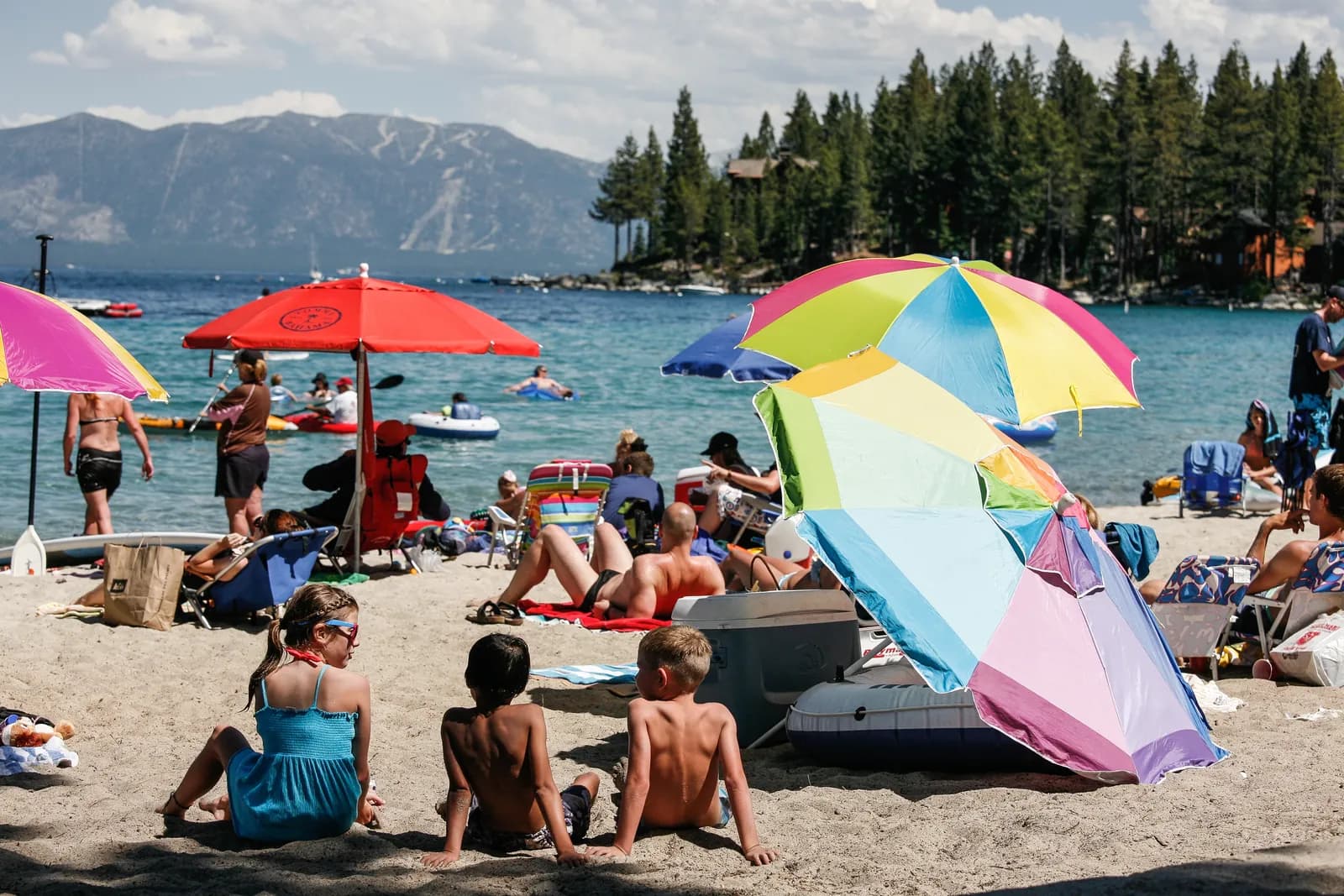 Beachgoers under bright umbrellas on a sandy South Lake Tahoe beach in July, with clear water and mountains across the lake
