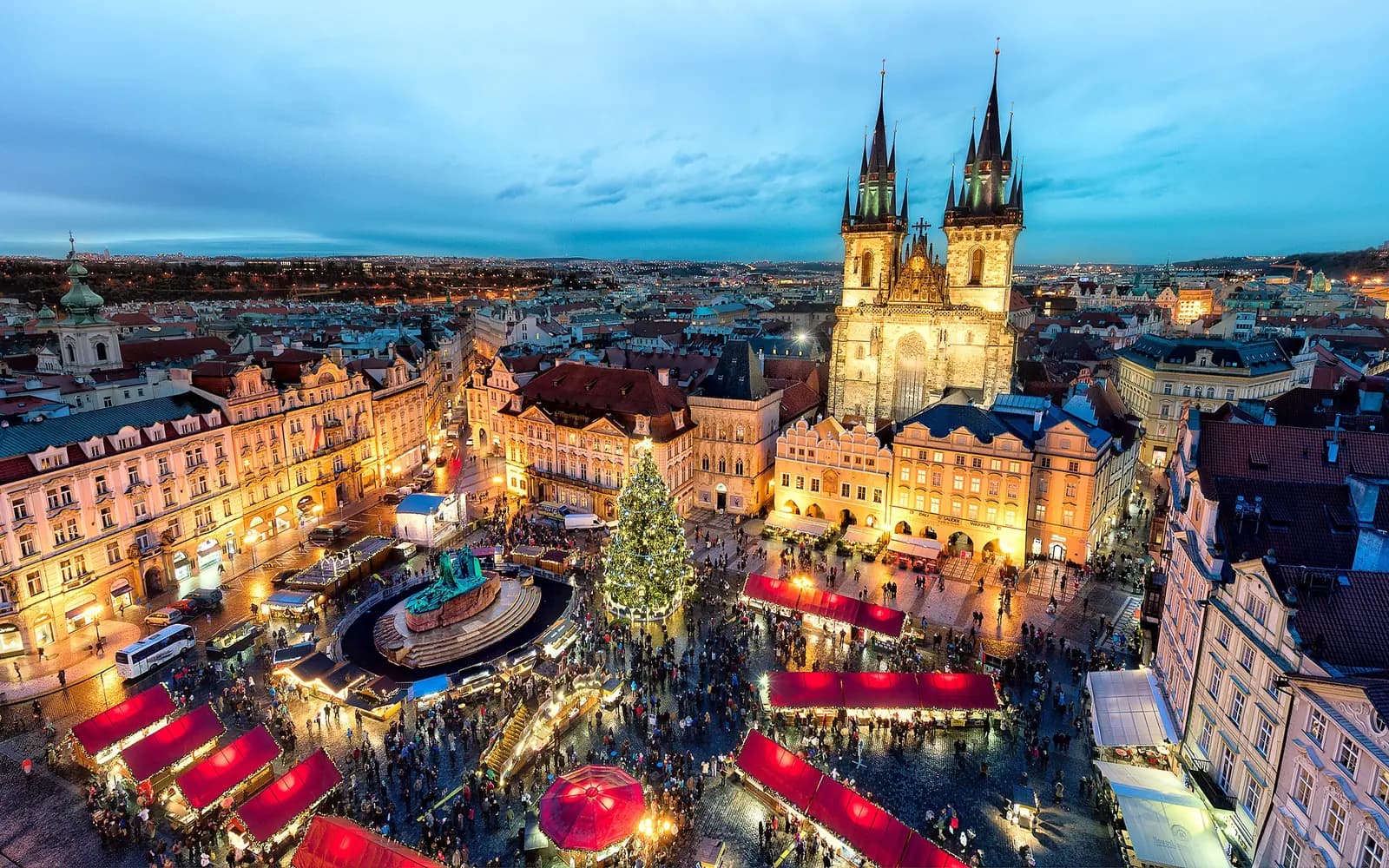 Prague Old Town Square Christmas market at dusk, with lit stalls, the tree and the Tyn Church towers at blue hour
