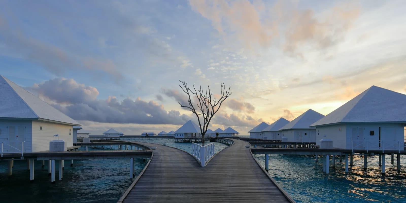 Wooden boardwalk curving between overwater villas above a Maldives lagoon at sunset