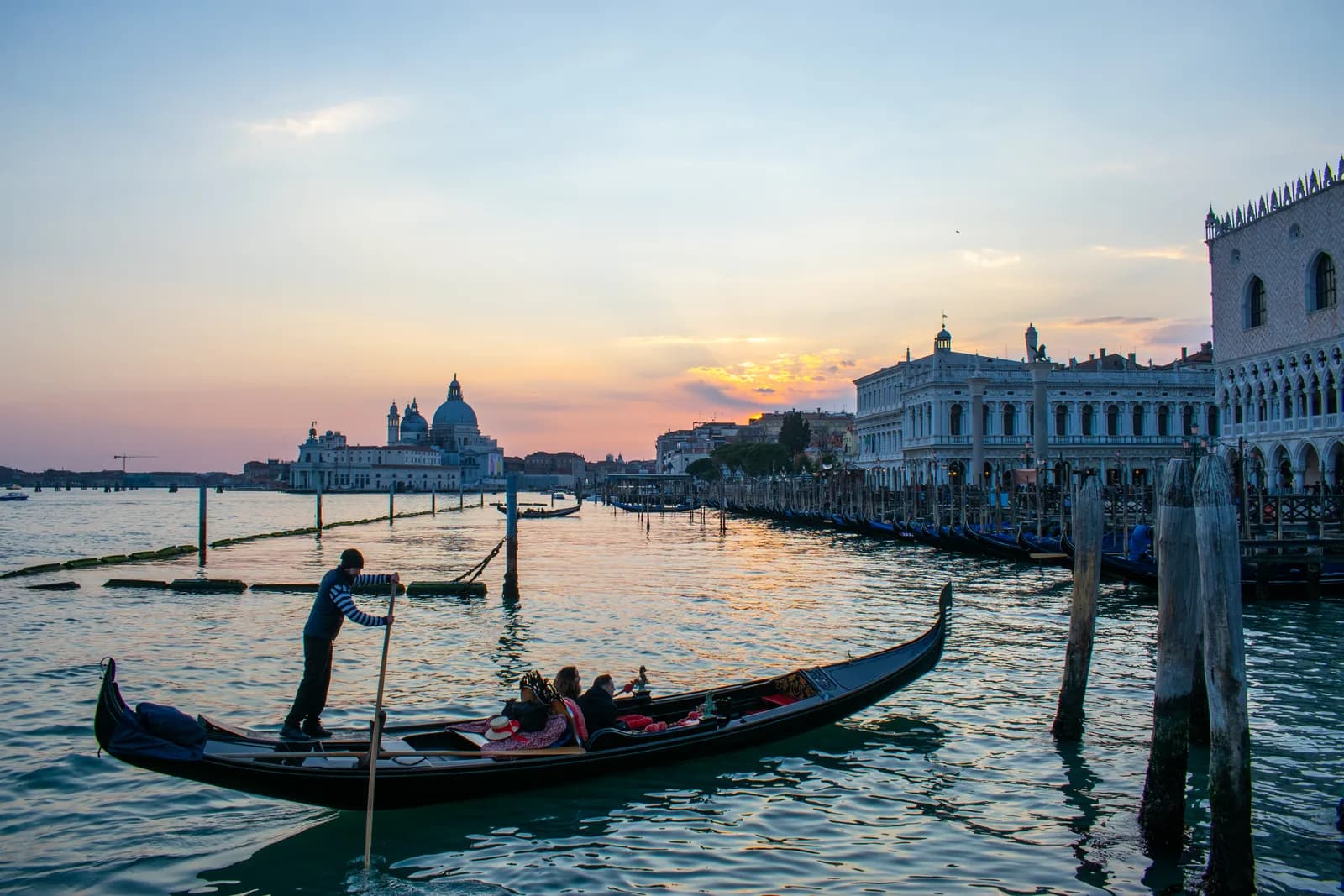 A gondolier steering a couple past the Doge's Palace at sunset in Venice, with Santa Maria della Salute on the horizon