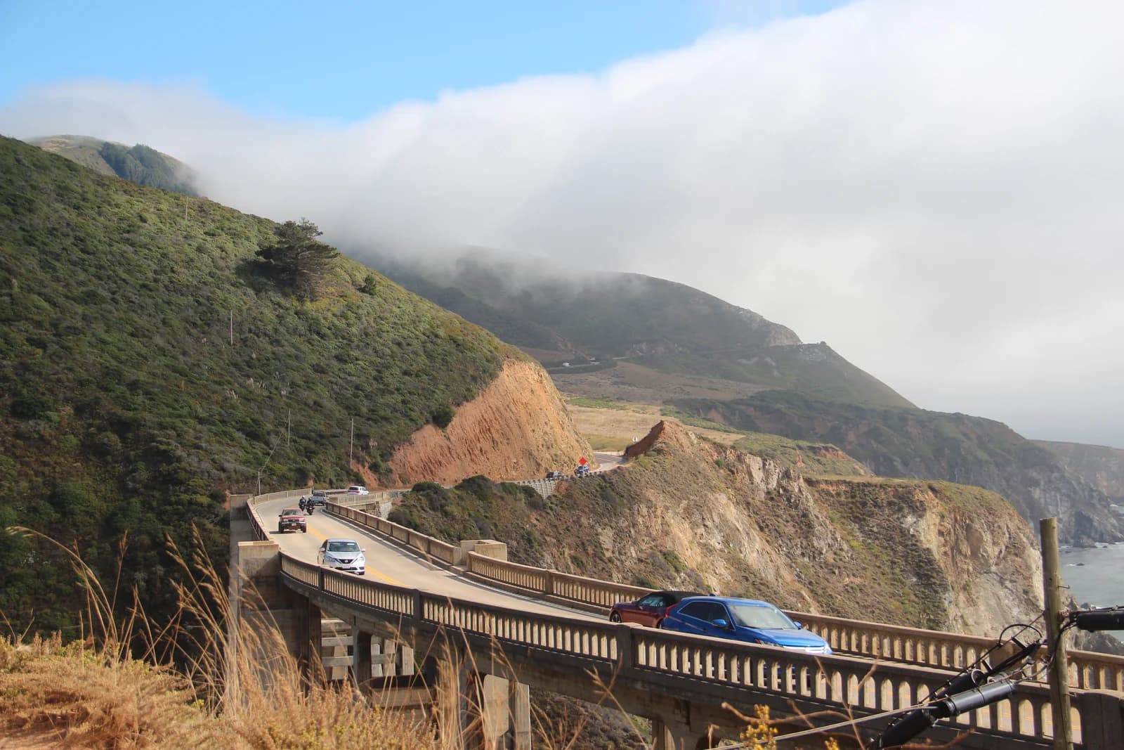 Cars cross Bixby Creek Bridge on Highway 1 in Big Sur, with a fog bank resting on the coastal hills behind