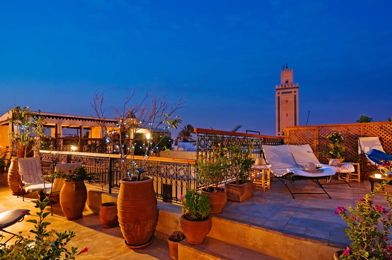 Riad rooftop terrace in the Marrakech medina at dusk, with sun loungers, potted plants and a lit minaret against the evening sky
