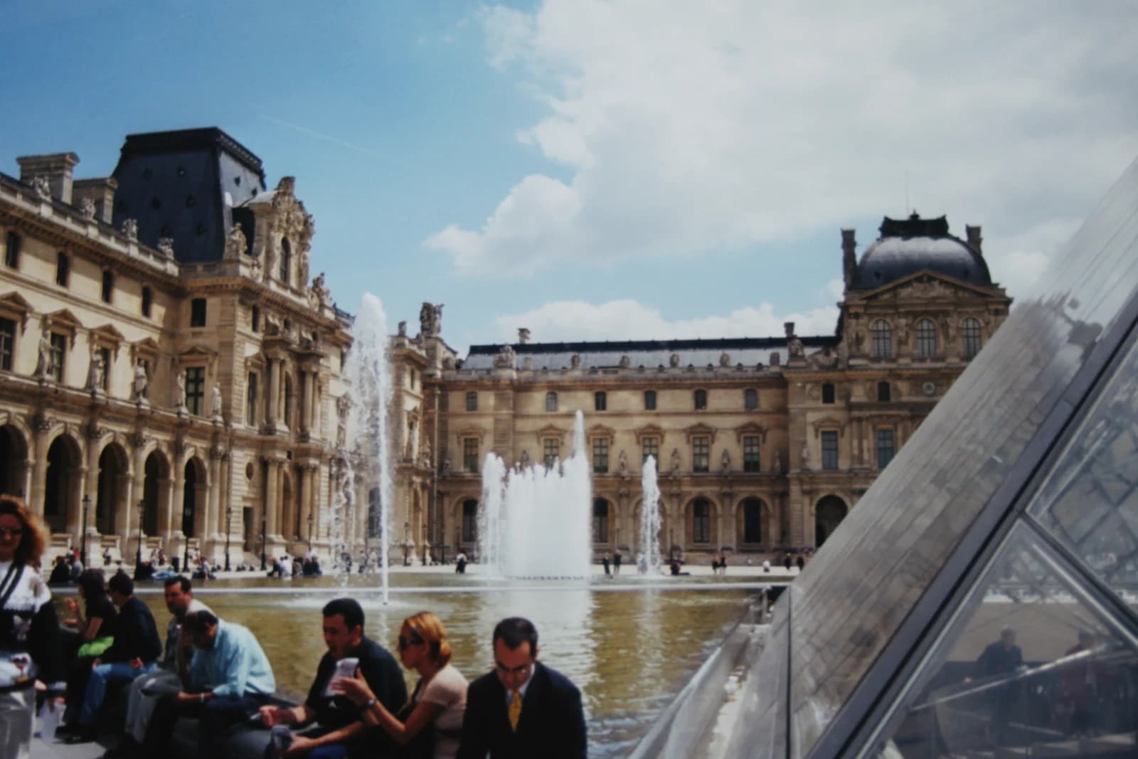 Visitors sitting beside the fountains in the Cour Napoleon at the Louvre, with the glass pyramid to one side