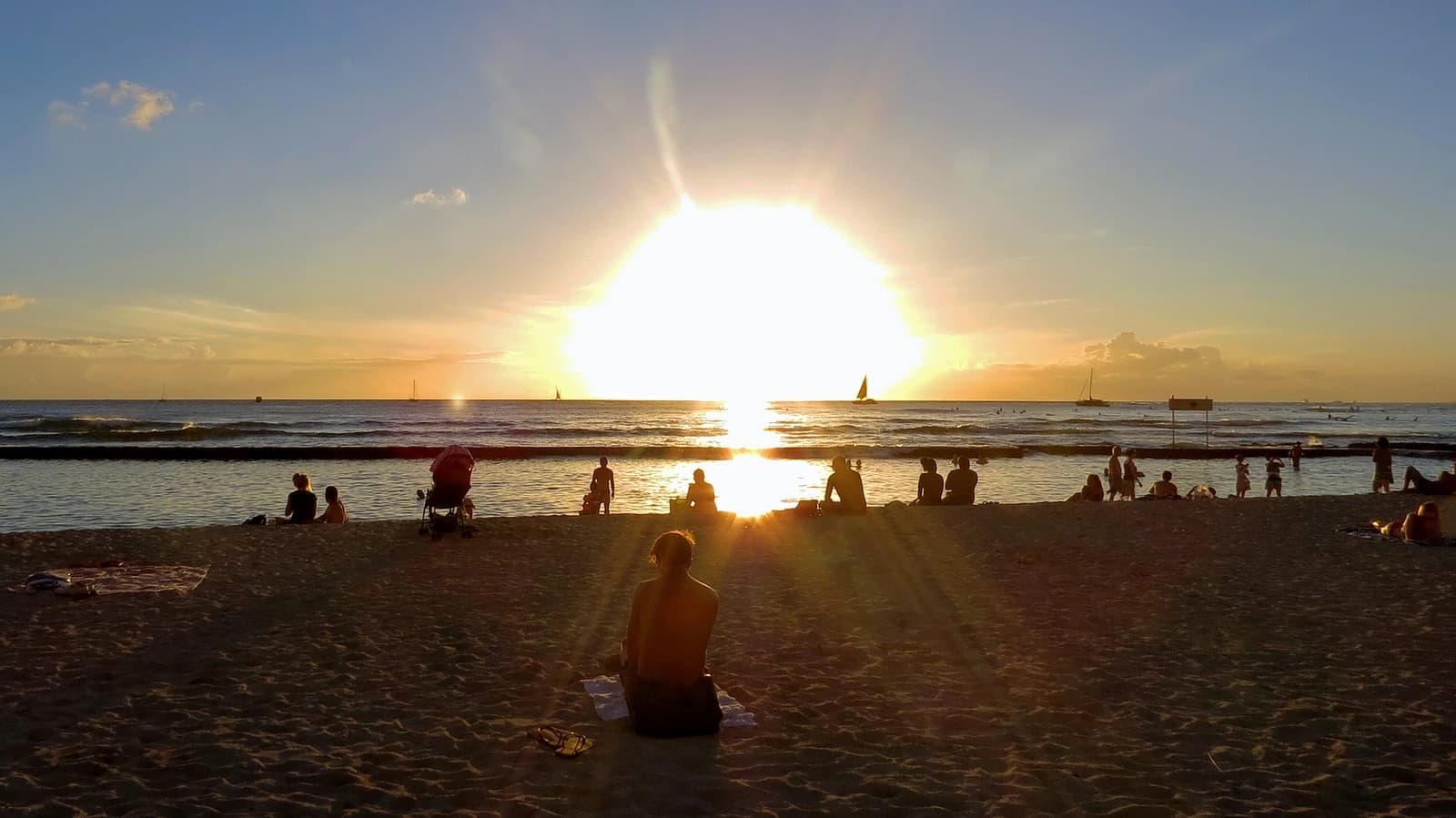 Sunset over Waikiki Beach, with families sitting on the sand and a low breakwater sheltering the water beyond