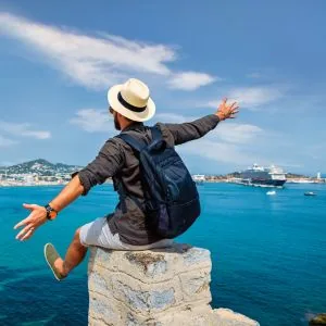 James, who writes the Journey Finder cruise guides, sitting on a rock above a harbour with a cruise ship anchored behind him